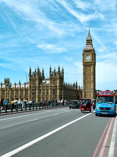 An image depicts a busy urban street scene in Westminster with the historic Elizabeth Tower, commonly known as Big Ben, and the Palace of Westminster prominent in the background under a clear blue sky with some wispy clouds. In the foreground, a road with multiple lanes is active with vehicles including a blue ice cream van, a black car, a small red auto rickshaw, and a dark-colored van, which are positioned near a curb. Several pedestrians are seen on the sidewalk, some walking and others standing, with additional people visible further along the street. Adjacent to the road, there is a pavement separated by a metal railing, and the scene suggests an area undergoing home or office removals, with furniture, cardboard boxes, or packing materials often involved in such relocations, although none are explicitly visible in this particular image. The image captures the transport and logistical aspect of moves in Westminster, supported by the presence of vehicles and the busy environment typical of home relocation and furniture transport activities, aligning with the theme of removals and moving services offered by companies like Removal Companies Westminster.