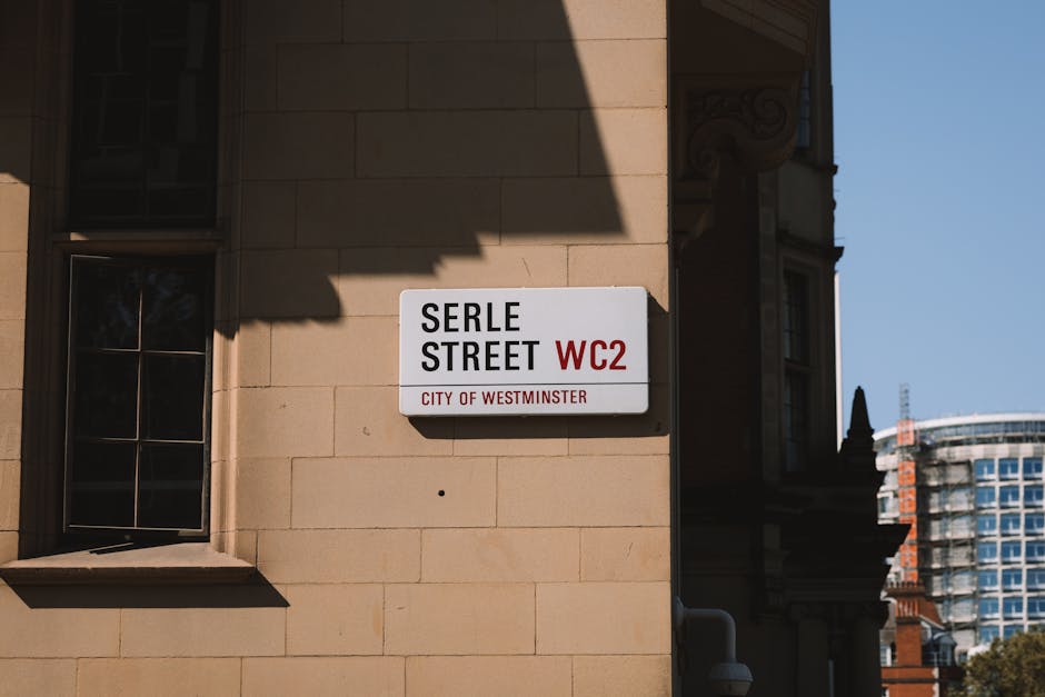 Close-up view of a street sign mounted on a beige stone building facade, indicating 'Serle Street WC2' in the City of Westminster. The sign has black and red text on a white background, with the words 'Serle Street' prominently displayed and 'WC2' in red, alongside a small red line separating it from the phrase 'City of Westminster' in red. Shadows cast by nearby structures partially obscure the building's window, which features a dark frame and reflective glass panes. In the background, part of a modern multi-storey building with glass windows and a spotted roofline is visible, set against a clear blue sky. The scene appears to be part of an urban environment where house removals and furniture transport services are performed, highlighting the area’s architectural style and street signage. The street sign's secure mounting and the surrounding environment suggest a typical location for moving logistics activities facilitated by removals companies like Westminster-based providers.