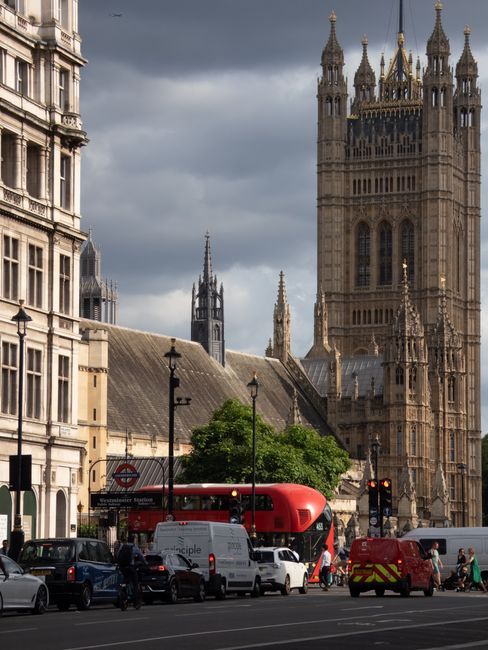 An image depicts a busy urban street scene in Westminster with the historic Elizabeth Tower, commonly known as Big Ben, and the Palace of Westminster prominent in the background under a clear blue sky with some wispy clouds. In the foreground, a road with multiple lanes is active with vehicles including a blue ice cream van, a black car, a small red auto rickshaw, and a dark-colored van, which are positioned near a curb. Several pedestrians are seen on the sidewalk, some walking and others standing, with additional people visible further along the street. Adjacent to the road, there is a pavement separated by a metal railing, and the scene suggests an area undergoing home or office removals, with furniture, cardboard boxes, or packing materials often involved in such relocations, although none are explicitly visible in this particular image. The image captures the transport and logistical aspect of moves in Westminster, supported by the presence of vehicles and the busy environment typical of home relocation and furniture transport activities, aligning with the theme of removals and moving services offered by companies like Removal Companies Westminster.