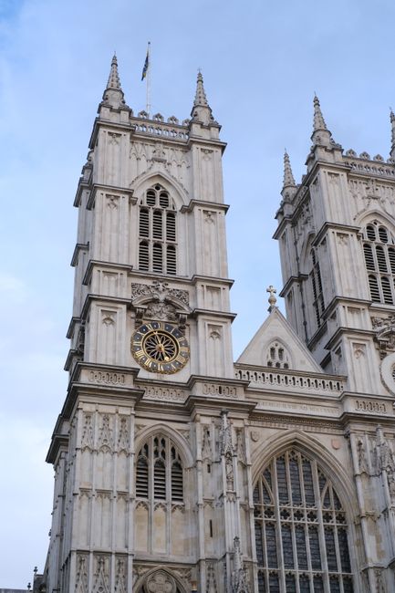 Exterior view of Westminster Abbey displaying two prominent stone towers with pointed spires and decorative gothic architectural details. The left tower features a large clock with a gold rim and black face, while the right tower has multiple narrow arched windows and intricate stone carvings. The sky is clear with a hint of light cloud cover. This image is associated with house removals and relocation services, highlighting the historic building as part of a move or logistics planning, showcasing the importance of careful handling and transportation of items near such landmark locations, with Removal Companies Westminster providing expert support for internal packing, furniture transport, and loading processes in close proximity to this renowned site.