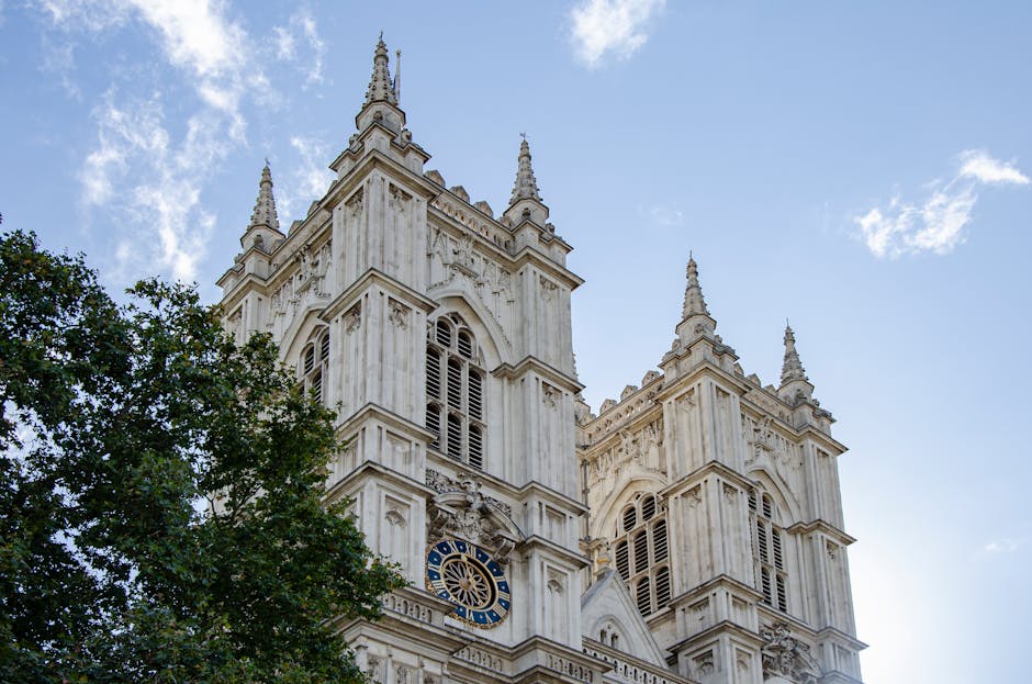 A close-up view of the upper section of Westminster Abbey’s white stone façade, showing detailed Gothic architectural features including spires, battlements, and arched windows with louvered shutters. The sky is clear with some scattered clouds, and green foliage partially obscures the lower part of the building. The image, associated with house removals and moving services, highlights the historic structure that may present narrow access and steps when undertaking a home relocation or furniture transport. This scene exemplifies the importance of careful planning and logistics handled by Removal Companies Westminster, especially considering the building's architecture and narrow access points during the loading and unloading process involved in professional removals.