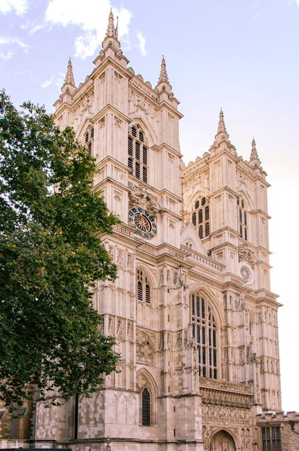 Exterior view of Westminster Abbey displaying two prominent stone towers with pointed spires and decorative gothic architectural details. The left tower features a large clock with a gold rim and black face, while the right tower has multiple narrow arched windows and intricate stone carvings. The sky is clear with a hint of light cloud cover. This image is associated with house removals and relocation services, highlighting the historic building as part of a move or logistics planning, showcasing the importance of careful handling and transportation of items near such landmark locations, with Removal Companies Westminster providing expert support for internal packing, furniture transport, and loading processes in close proximity to this renowned site.